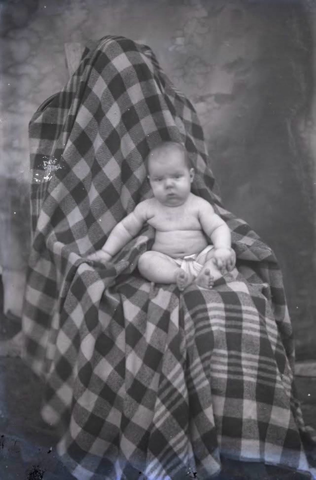 A baby sits on a chair draped in a large, black-and-white checkered blanket, with a person hidden beneath the fabric behind the child.