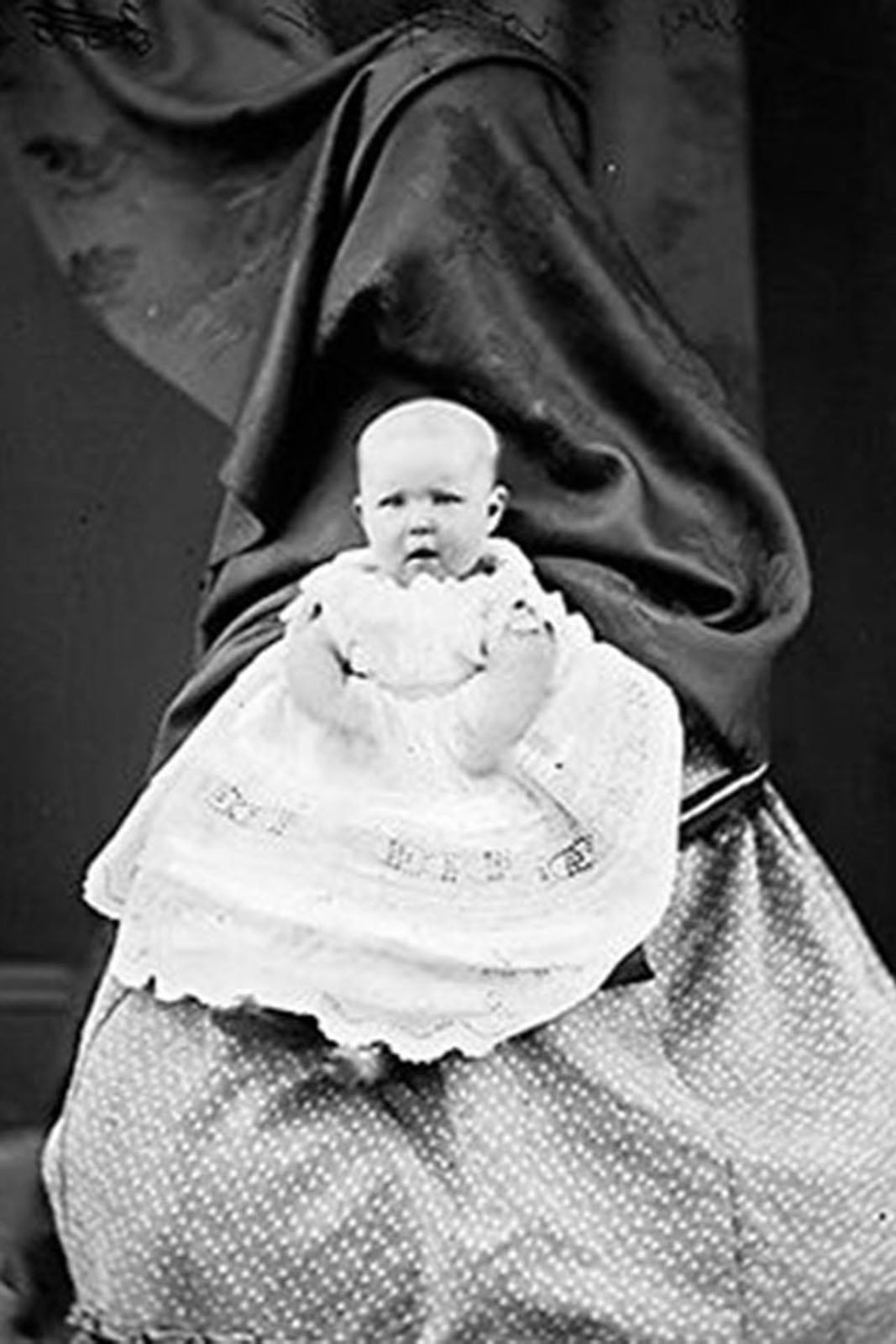 A baby in a white lace dress sits on the lap of a person completely obscured by a dark, draped fabric.