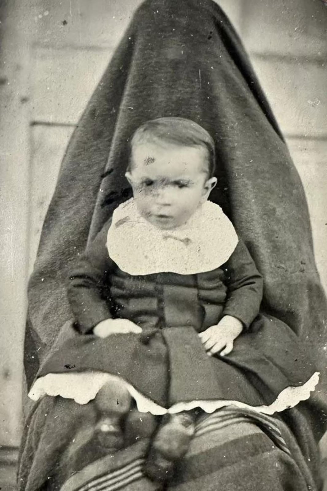 A vintage black-and-white portrait of a baby sitting on the lap of a person completely covered by a dark, hooded fabric shroud.