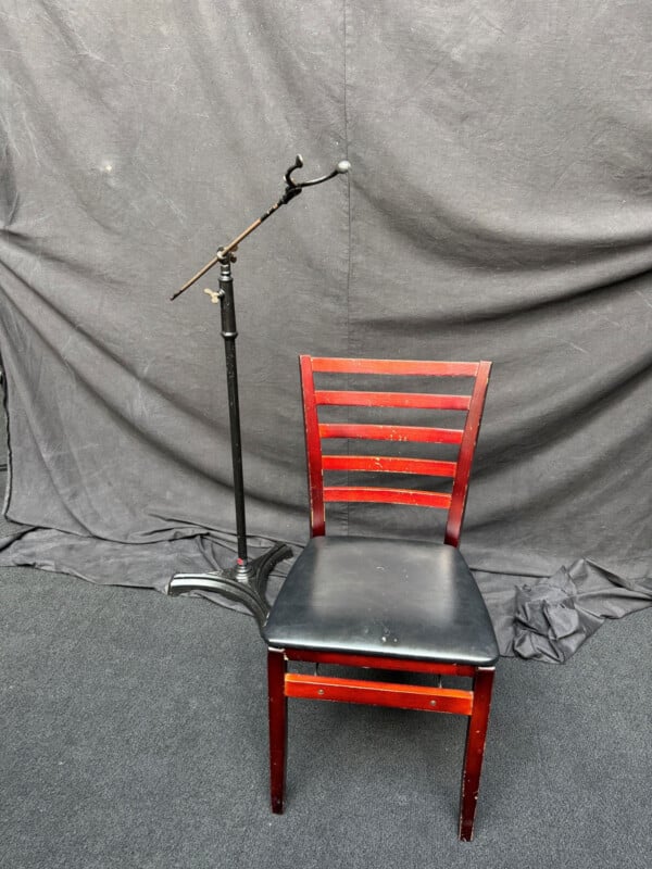 A black metal stand with an adjustable arm sits next to a wooden chair with a black padded seat and a ladder-back design against a dark fabric backdrop.